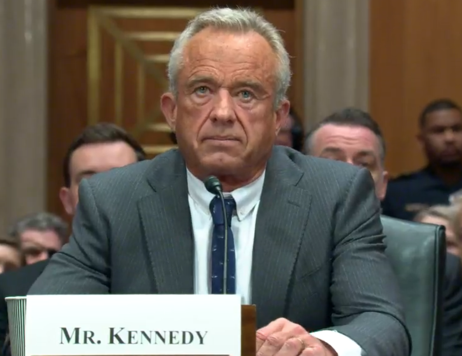 AI-generated neutral companion image — Senate hearing room with name placards and microphones; audience and press present; illustrative